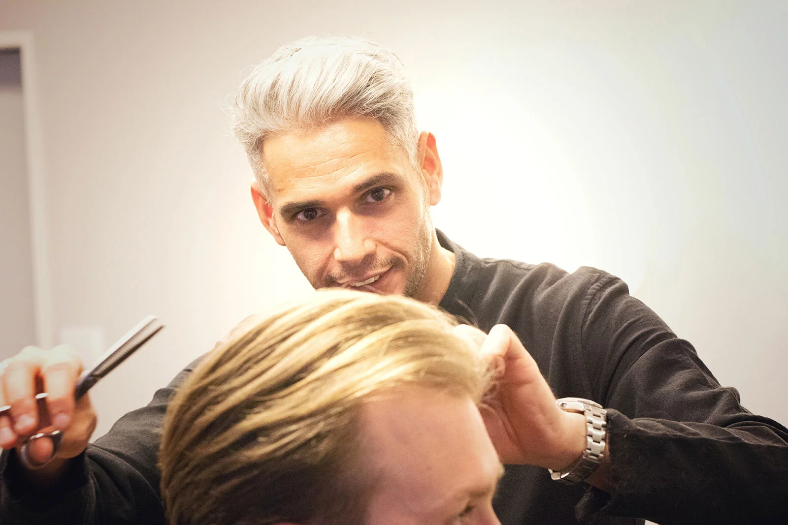 Mo schneidet einem Kunden die Haare und schaut dabei im Spiegel in die Kamera